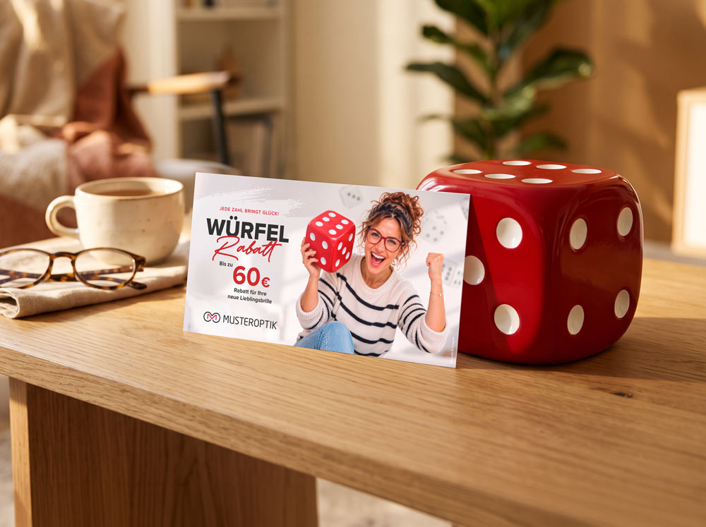 Würfel-Rabatt – Szene Motiv Frau Maxipostkarte „Würfel-Rabatt“ mit Motiv Frau, an einen großen roten Würfel gelehnt, auf einem Holztisch im Wohnzimmer.